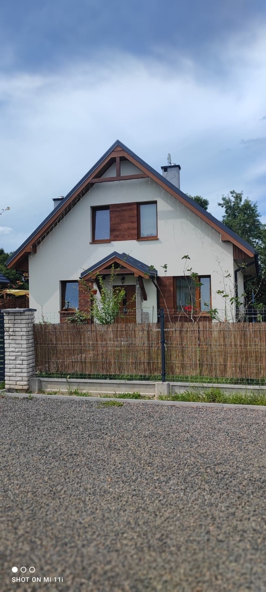 Dwukondygnacyjny dom jednorodzinny z jasną elewacją, ciemnym dachem i drewnianymi elementami wykończeniowymi okien, otoczony płotem z siatki i maty bambusowej, oświetlony girlandą lampek.