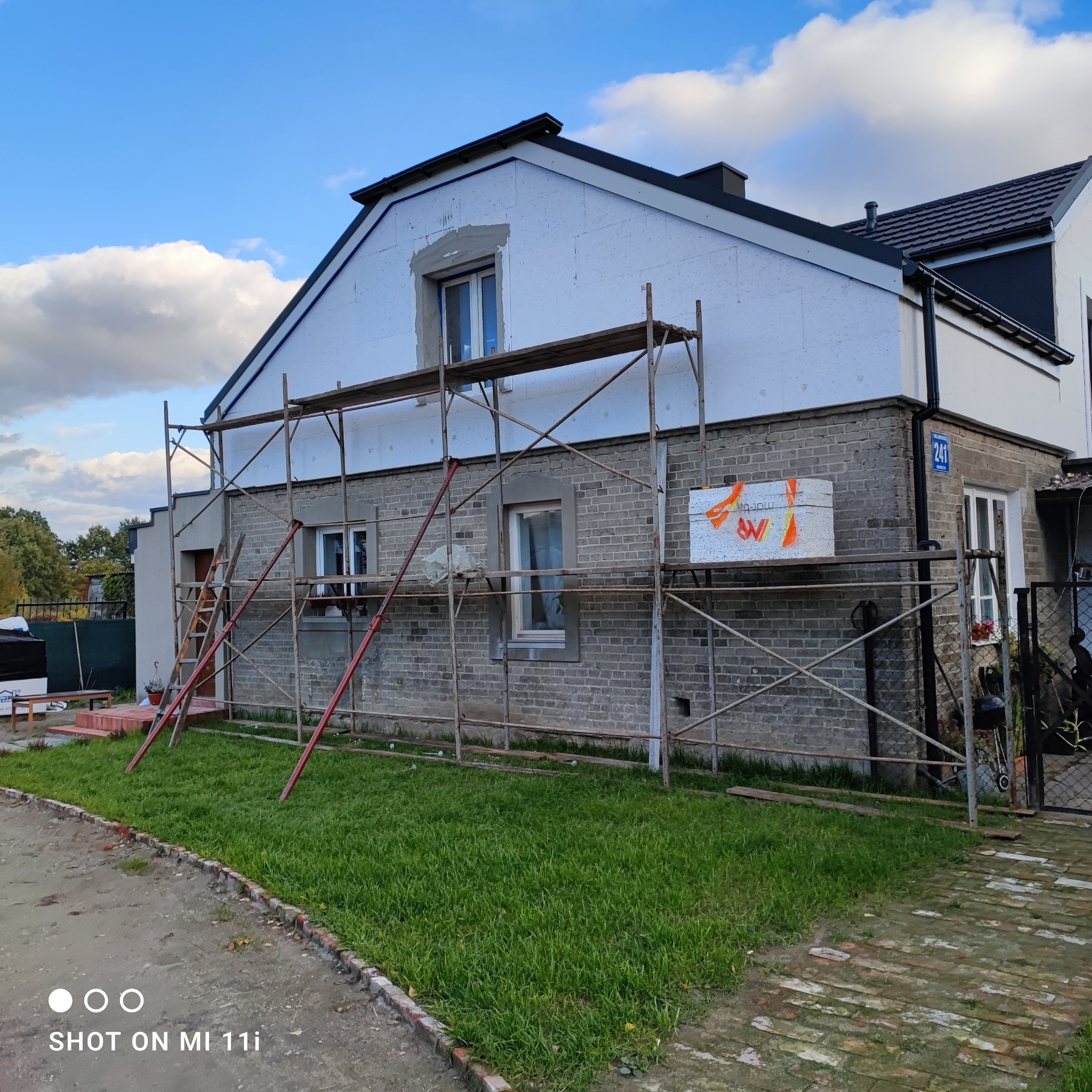 Dom jednorodzinny w trakcie ocieplania, widoczne rusztowania, styropian na elewacji, częściowo otynkowane okna, numer domu 241.