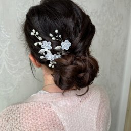 ZmalowAna by Anna Prażmo - Upięcie włosów w kok z ozdobami kwiatowymi i perłami, widok z tyłu na tle jasnej ściany z delikatnym wzorem. Subtelna elegancja.
