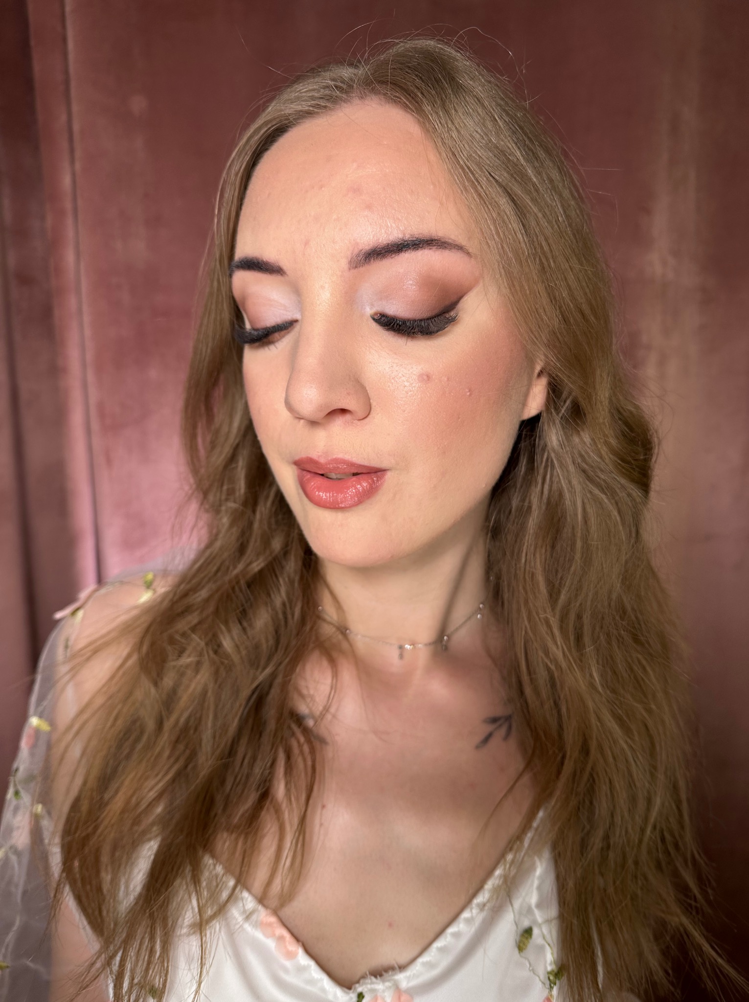 Portret kobiety z zamkniętymi oczami prezentującej delikatny makijaż w odcieniach różu i brązu, z widocznym tatuażem na szyi i subtelną biżuterią.