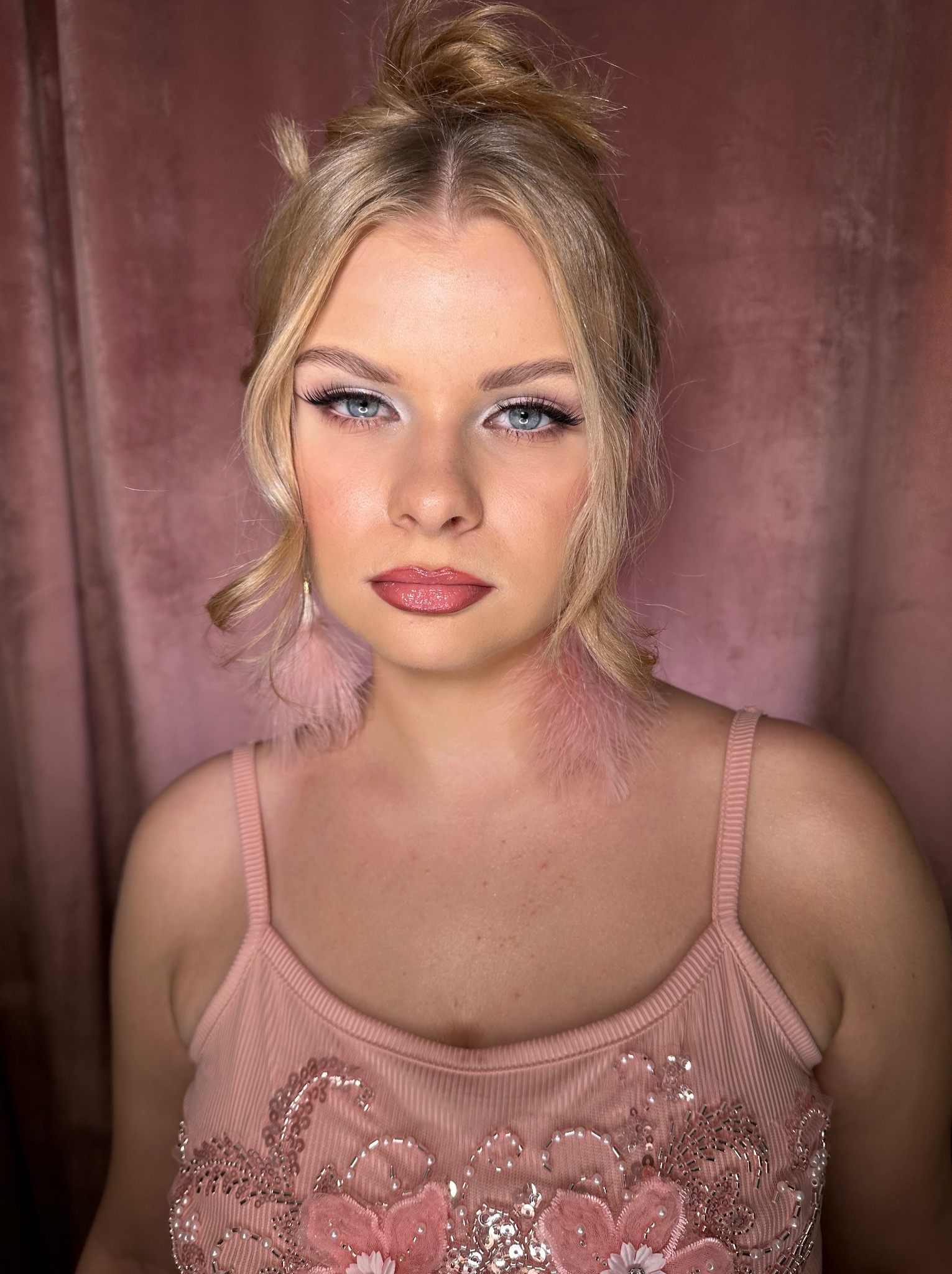 Portret kobiety z blond włosami upiętymi w kok, w różowej sukience z ozdobami, z delikatnym makijażem i kolczykami z piór na tle różowej zasłony.