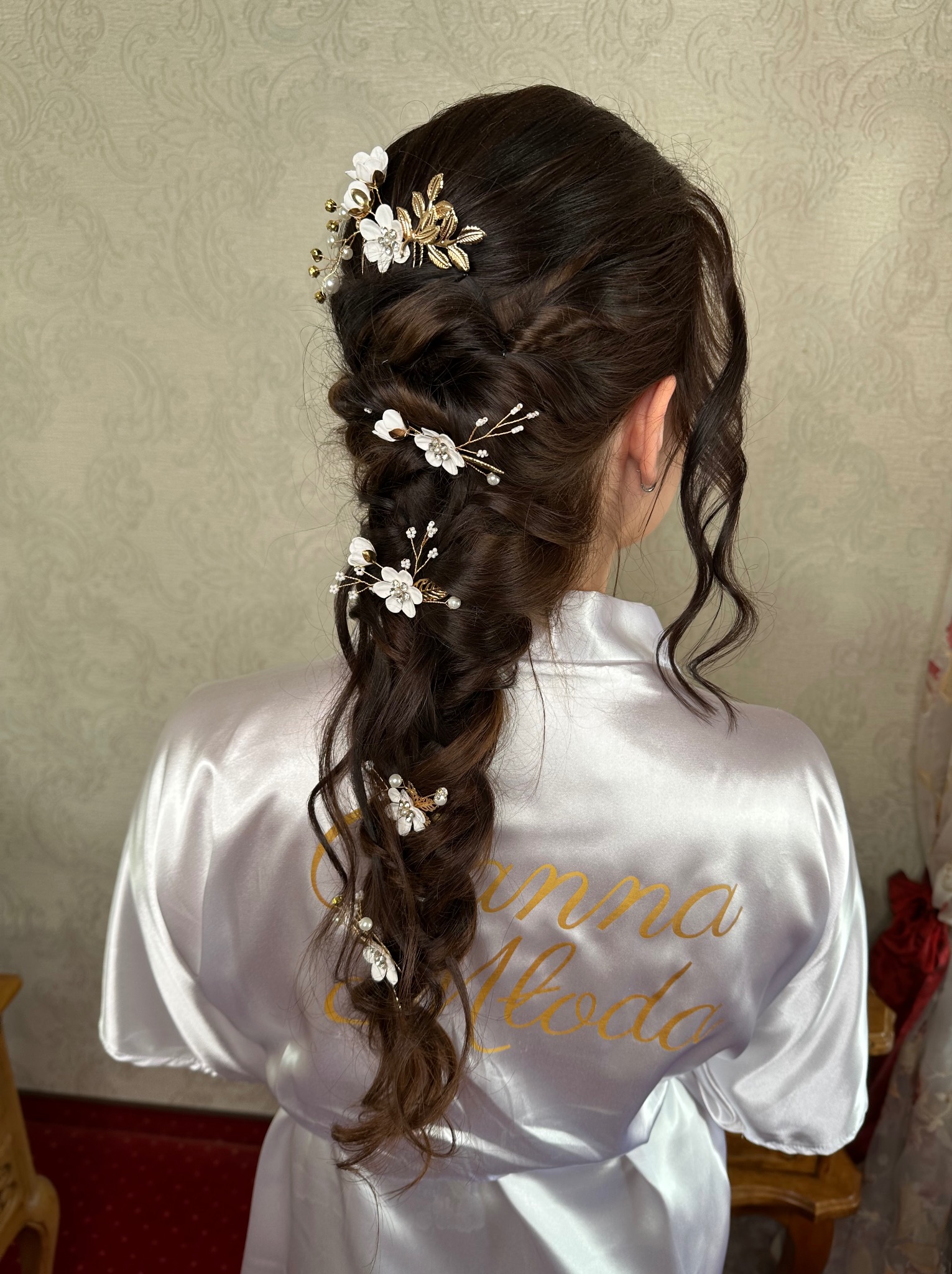 Upięcie ślubne z warkoczem i kwiatowymi ozdobami, widok z tyłu, na ramionach biały szlafrok z napisem 'Panna Młoda'.