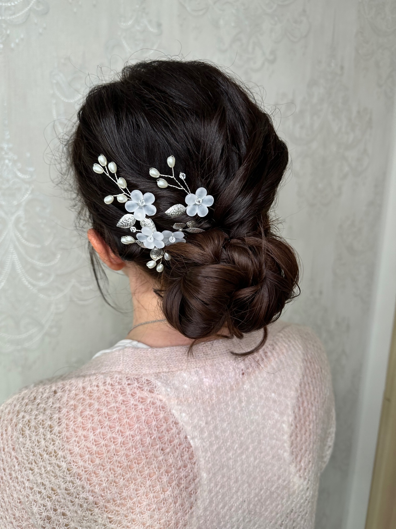 Upięcie włosów w kok z ozdobami kwiatowymi i perłami, widok z tyłu na tle jasnej ściany z delikatnym wzorem. Subtelna elegancja.
