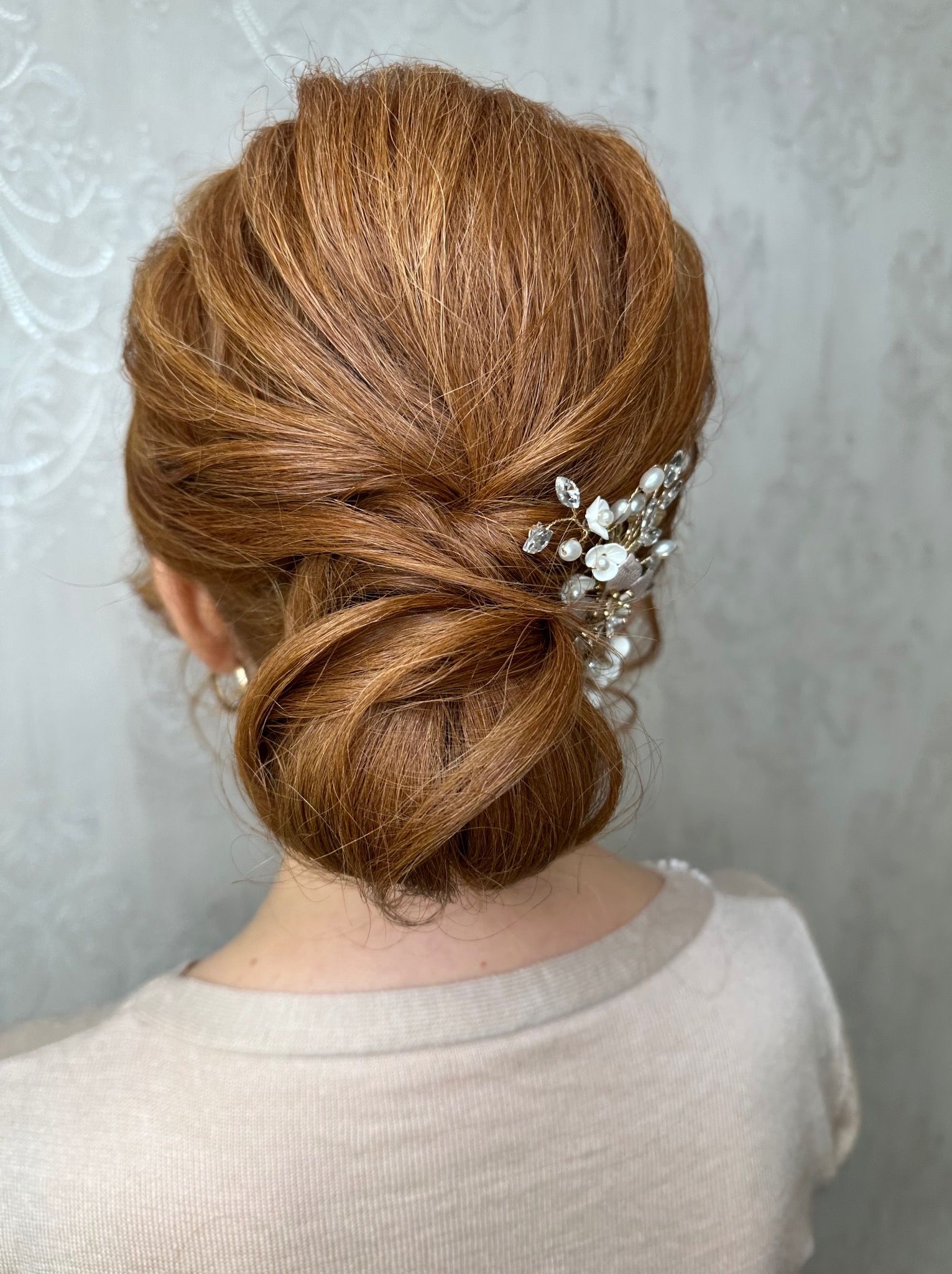Upięcie ślubne z rudych włosów z ozdobną spinką z białymi kwiatami i perłami, widok z tyłu na tle jasnej ściany. Elegancka fryzura.