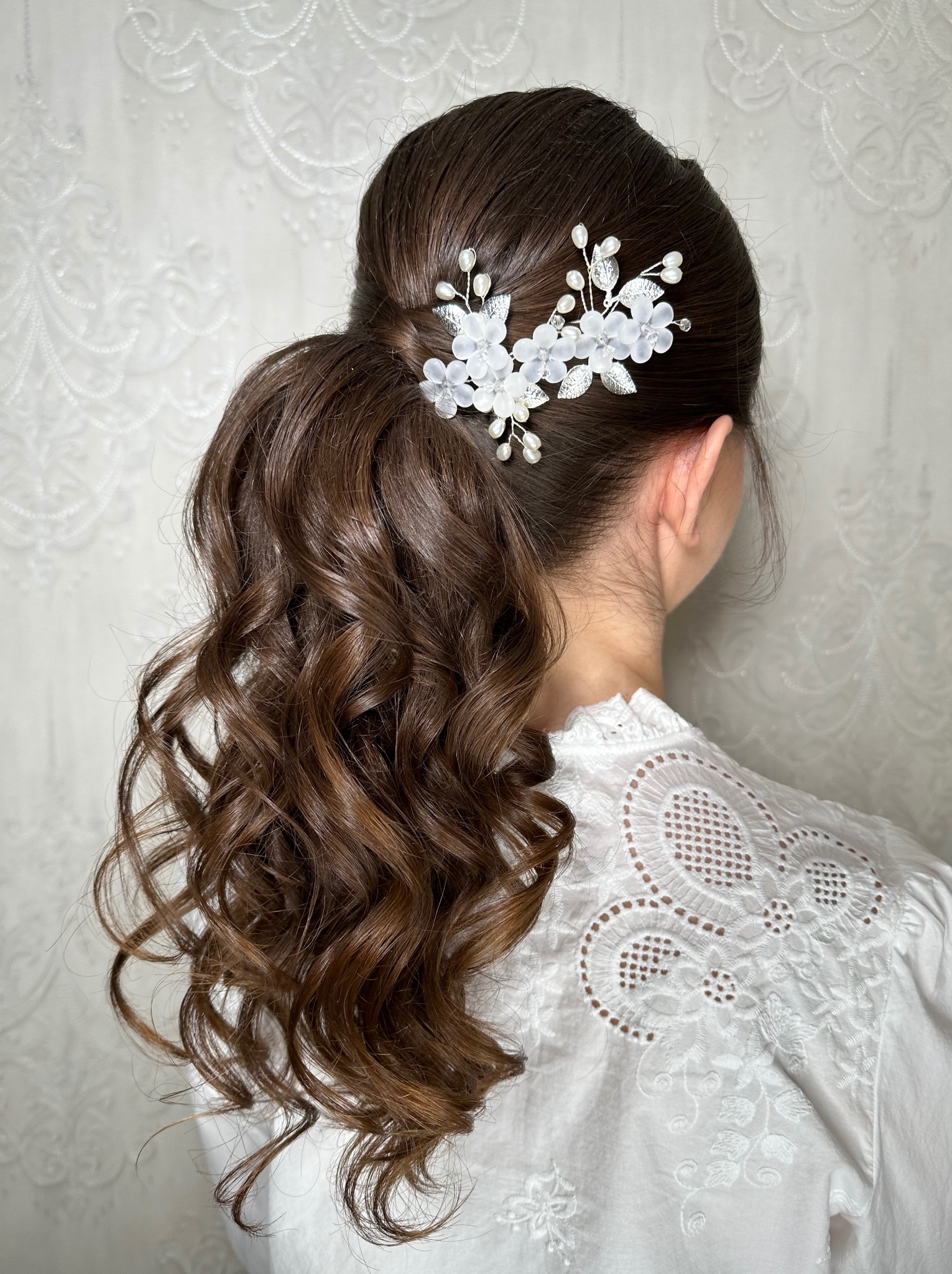 Upięcie włosów w niski kucyk z ozdobną spinką z białych kwiatów i srebrnych listków na tle białej, wzorzystej tapety i ażurowej bluzki.