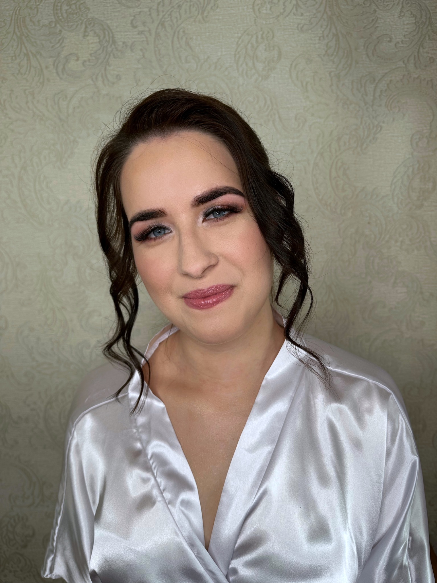 Portret kobiety z delikatnym makijażem ślubnym: różowe cienie, podkreślone rzęsy i błyszczące usta, w satynowym szlafroku na tle wzorzystej tapety.