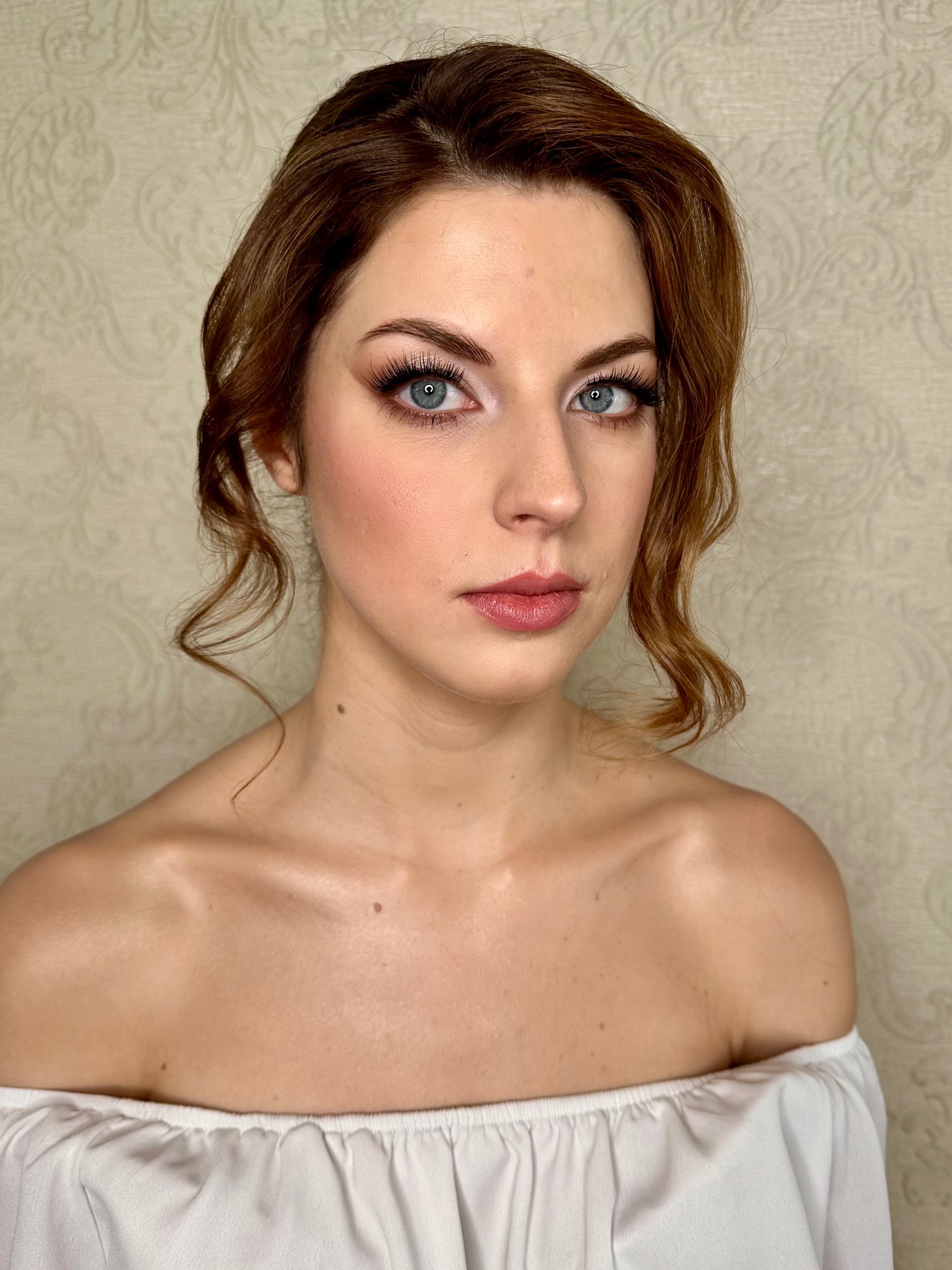 Portret kobiety z delikatnym makijażem i upiętymi włosami. Uwagę przyciągają podkreślone oczy i subtelny odcień ust. Tło stanowi ściana w jasnym, stonowanym kolorze.