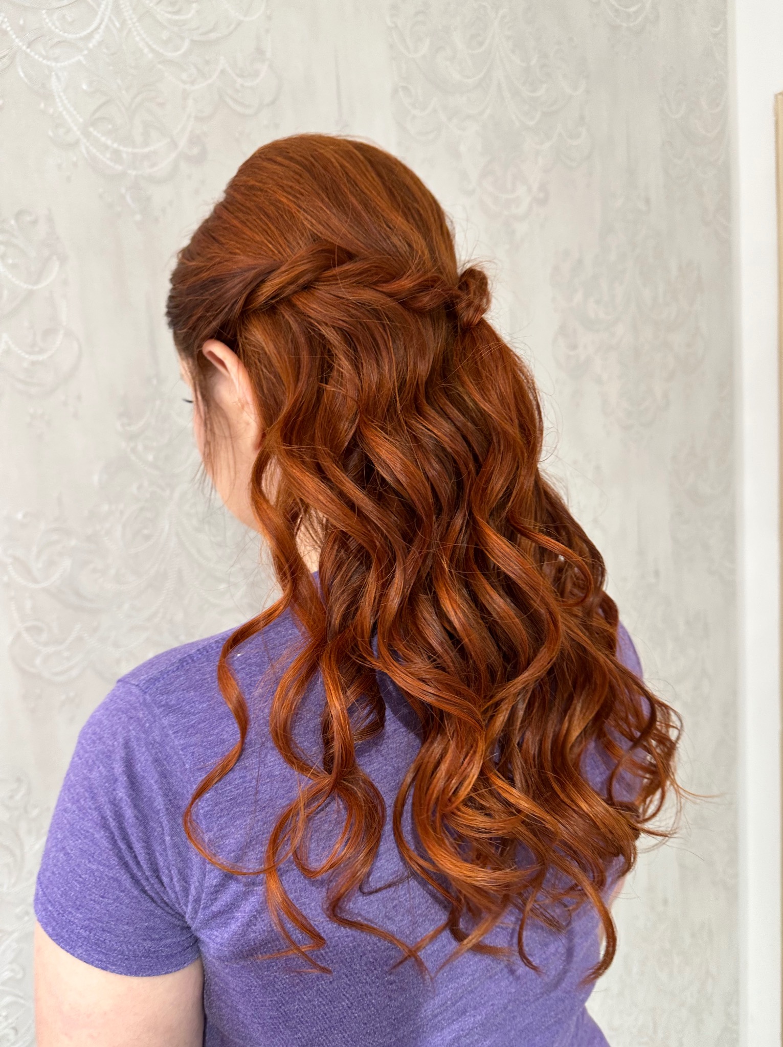 Rude włosy upięte w romantyczną fryzurę z warkoczem i lokami, widok z tyłu na tle jasnej ściany. Subtelne upięcie na specjalną okazję.