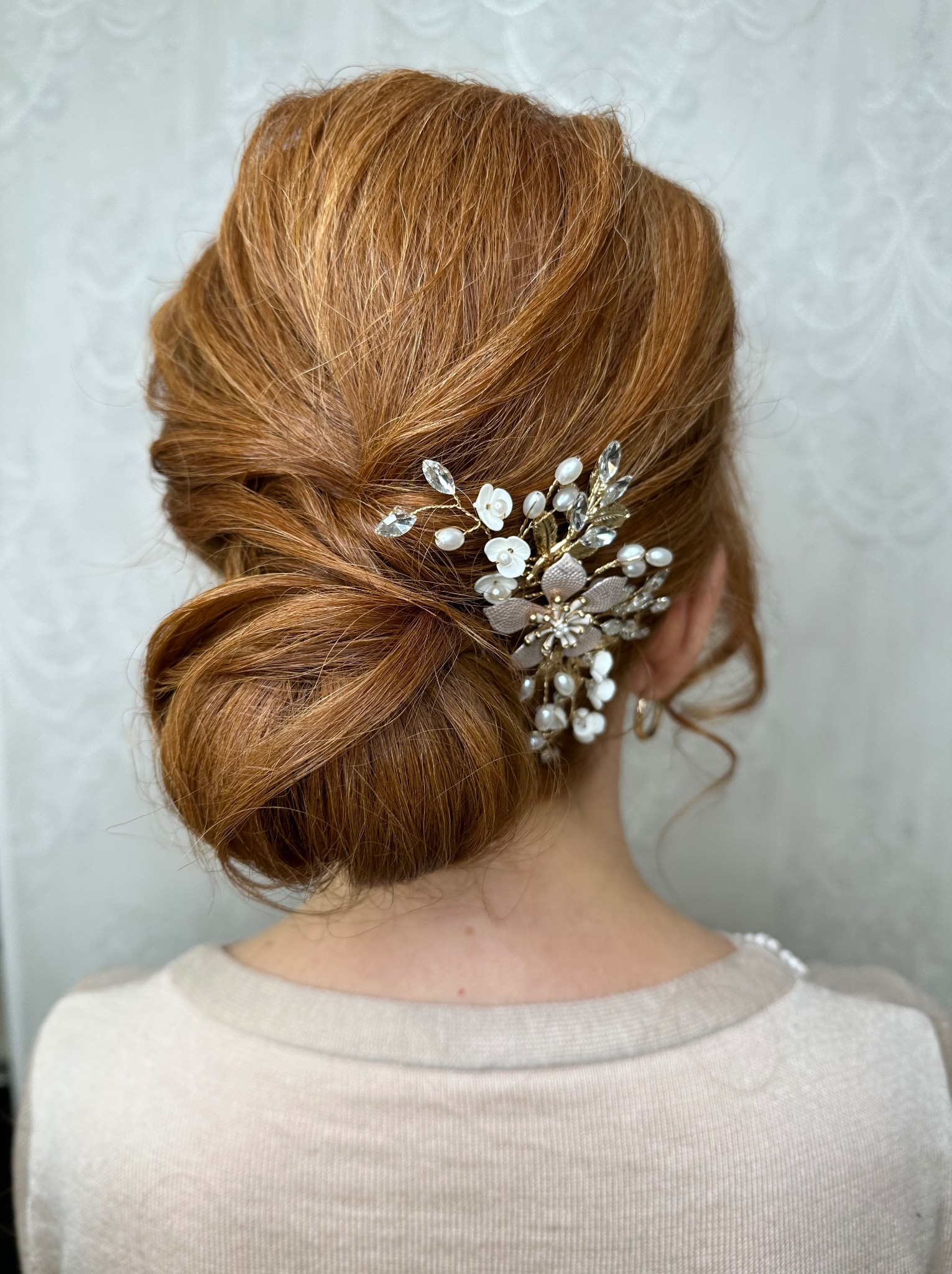 Upięcie ślubne z rudych włosów z ozdobną spinką z perełkami i kryształkami, widok z tyłu na jasnym tle. Elegancka fryzura na specjalną okazję.