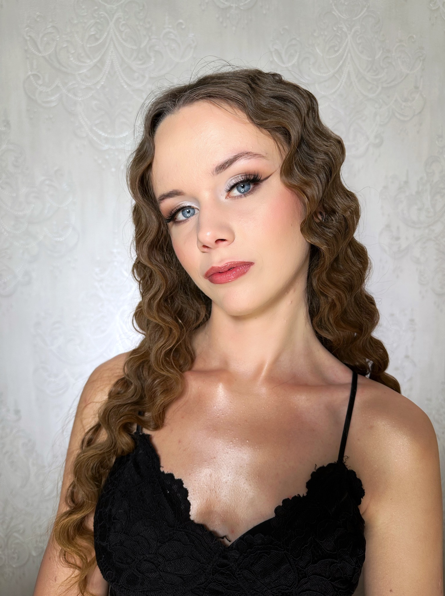 Portret kobiety z profesjonalnym makijażem oczu i ust, oraz z lokami w stylu retro na tle ściany z delikatnym wzorem. Ujęcie od przodu, oświetlenie studyjne.