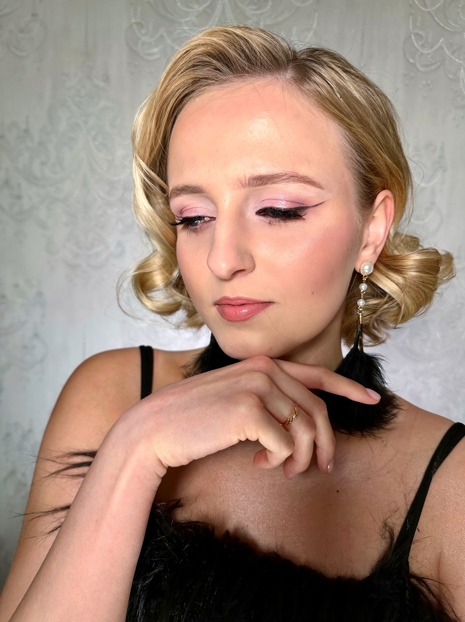 Portret kobiety z blond włosami w stylu lat 20. z wyrazistym makijażem oczu i ust, oraz eleganckimi kolczykami z piórami. Dyskretny wzór na ścianie w tle.