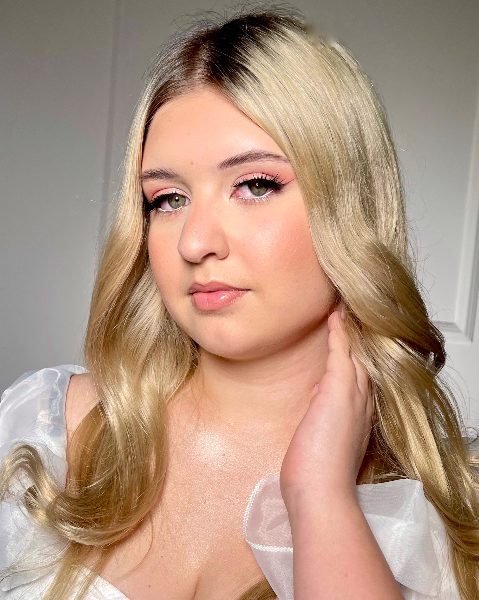 Portret blondynki z delikatnym, różowym makijażem oczu i błyszczącymi ustami, dłoń przy szyi, w bluzce z prześwitującego materiału, na jasnym tle.