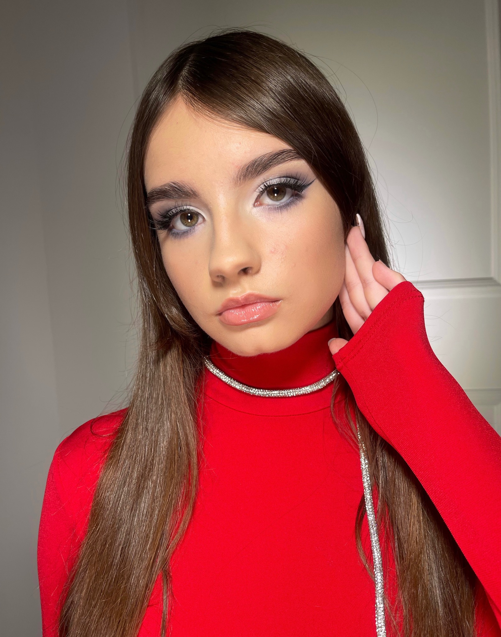 Młoda brunetka z pełnym makijażem oczu w odcieniach szarości i błyszczącymi ustami, ubrana w czerwoną bluzkę z golfem i srebrny naszyjnik.