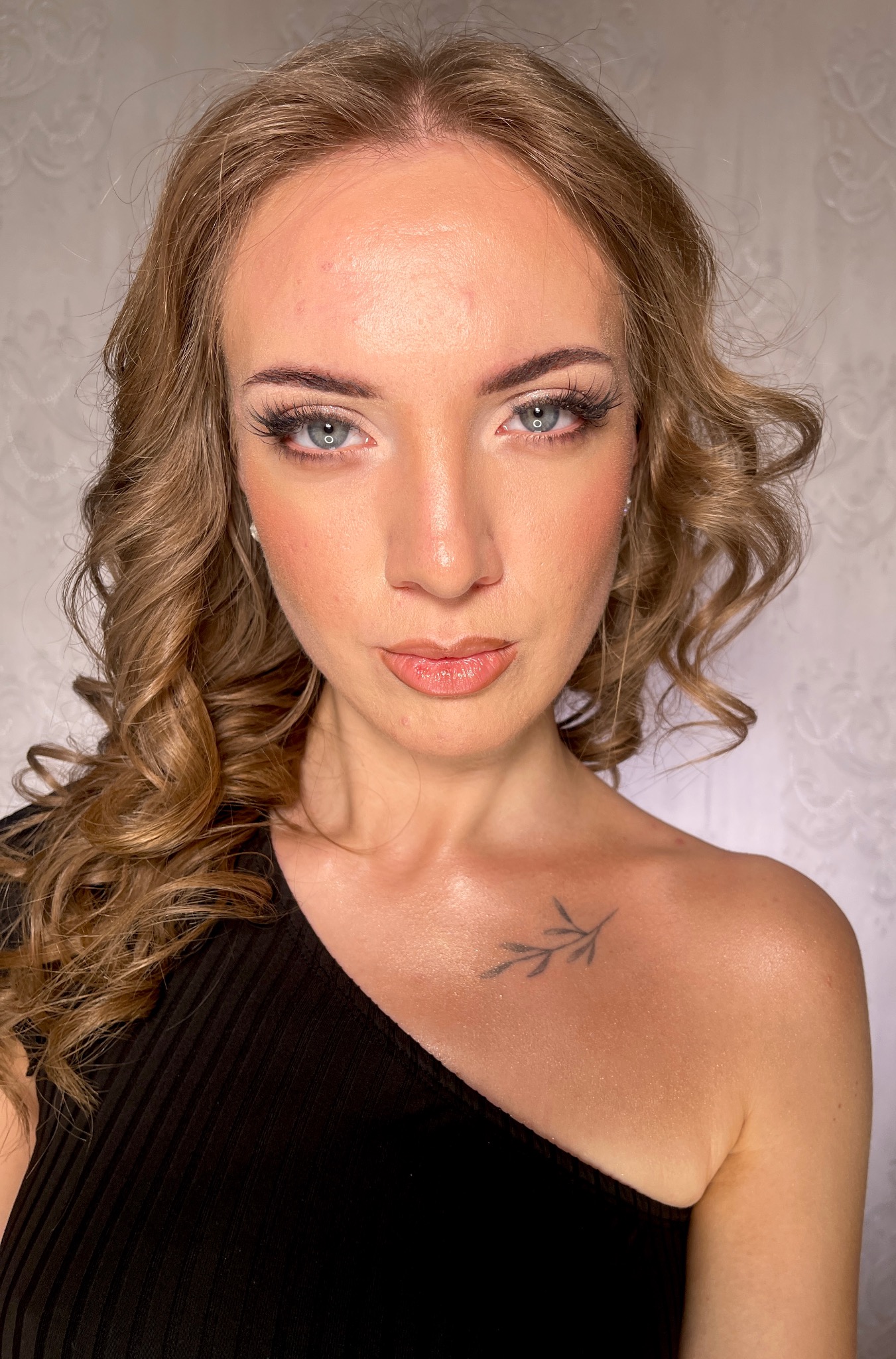 Portret kobiety z profesjonalnym makijażem i fryzurą, widoczny tatuaż na ramieniu, delikatne fale, podkreślone oczy i usta w odcieniach nude. Elegancka stylizacja.