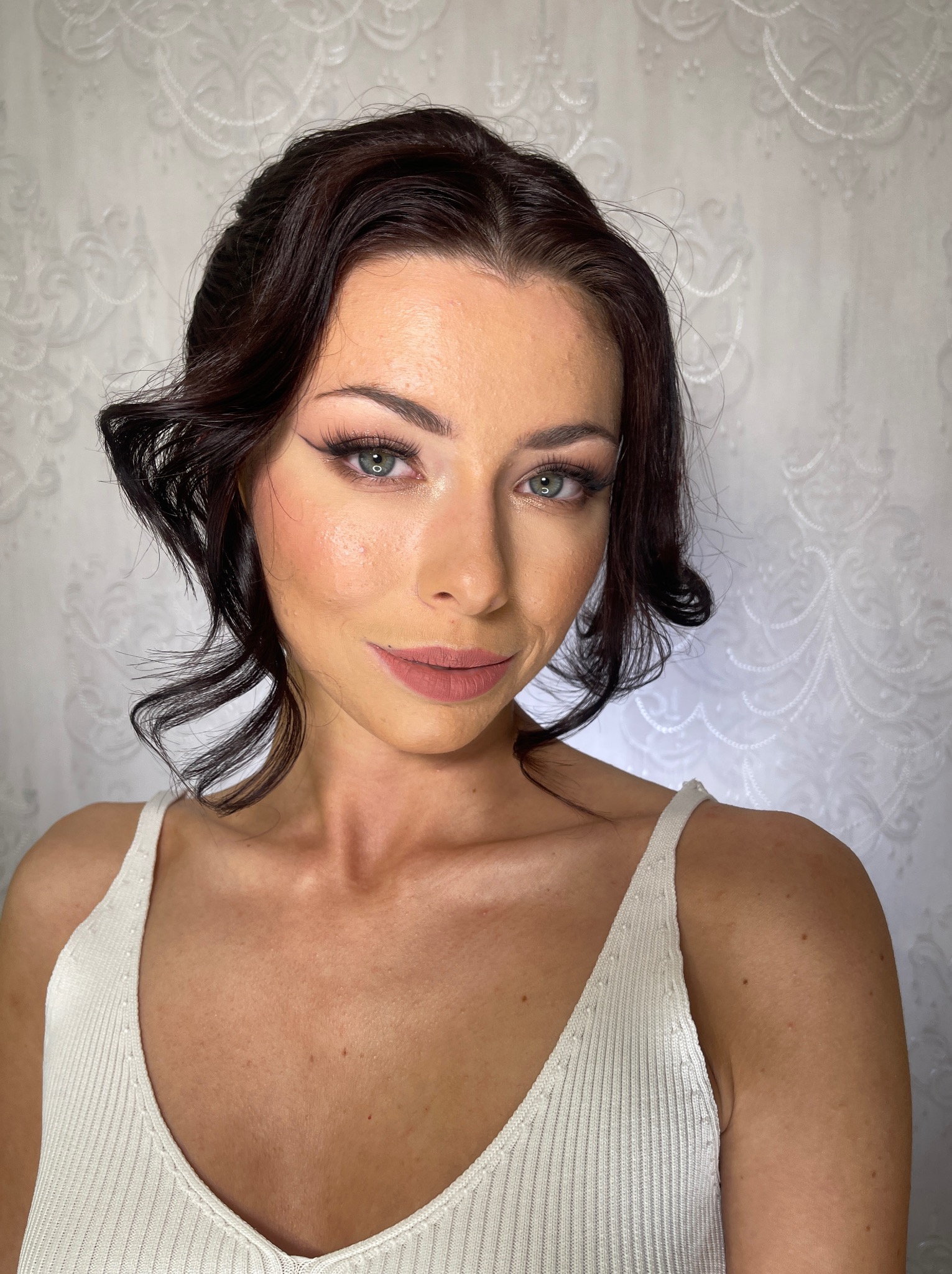 Portret kobiety z profesjonalnym makijażem i upiętymi, ciemnymi włosami na tle wzorzystej ściany. Widoczne delikatne podkreślenie oczu i ust.