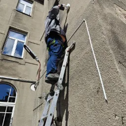 Pracownik na drabinie montuje białą kamerę na szarej, chropowatej ścianie budynku, widoczne niebieskie spodnie robocze i fragment lampy ulicznej.