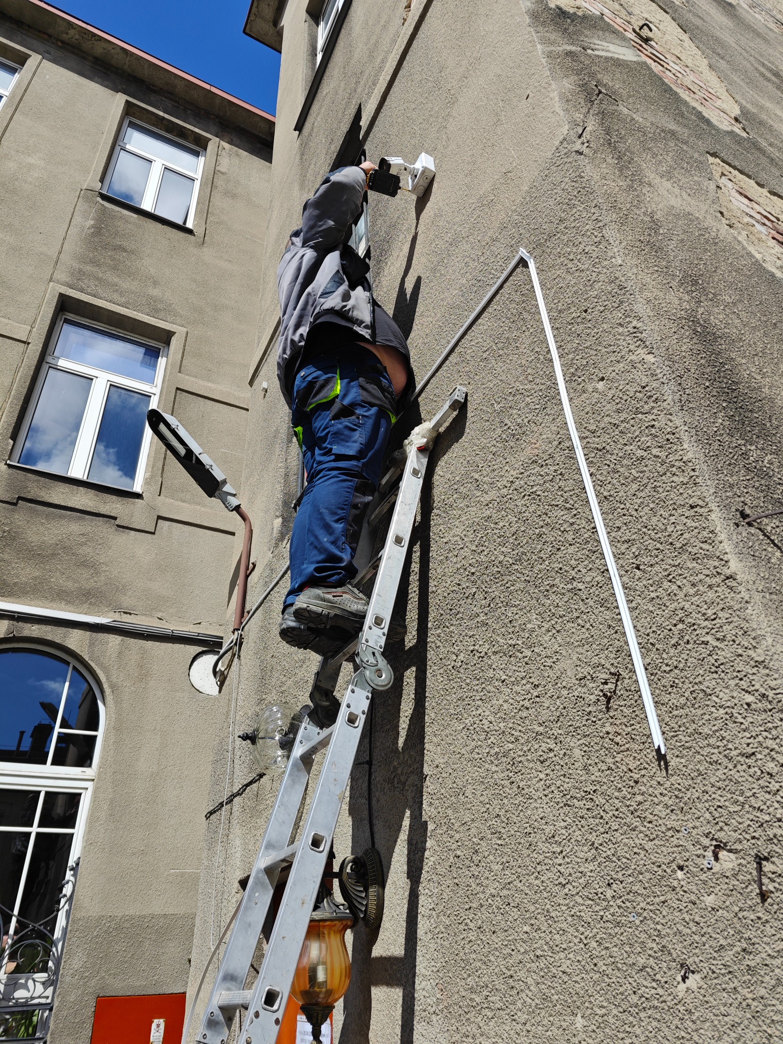 Pracownik na drabinie montuje białą kamerę na szarej, chropowatej ścianie budynku, widoczne niebieskie spodnie robocze i fragment lampy ulicznej.