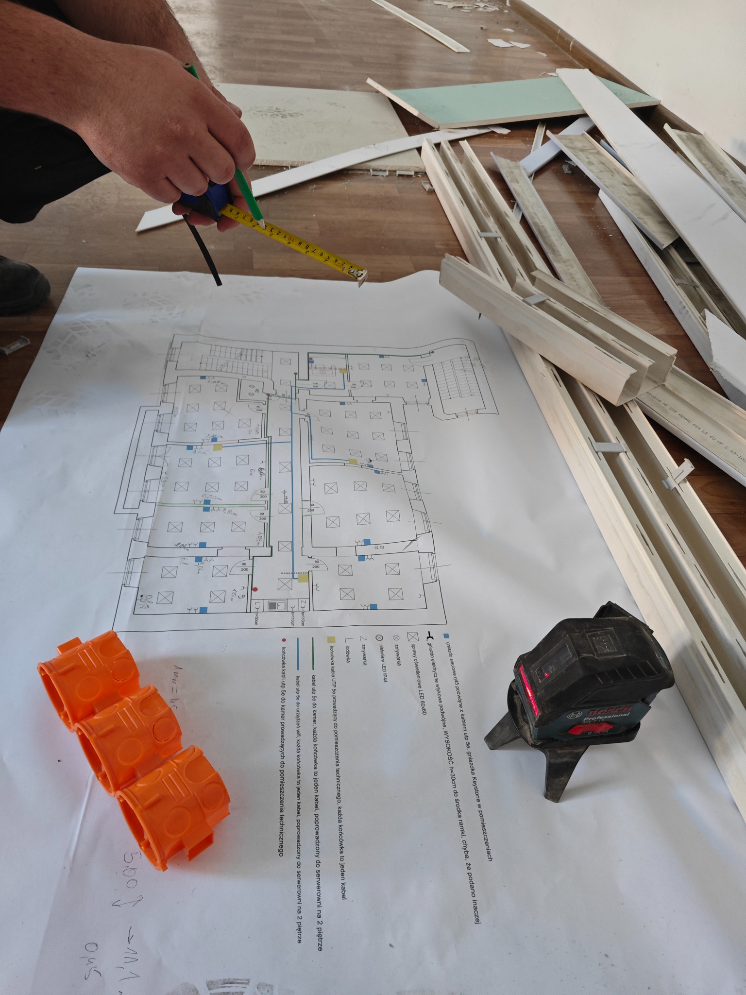 Pomiar odległości na planie instalacji elektrycznej za pomocą miarki i ołówka, obok puszki instalacyjne, poziomica laserowa i elementy wykończeniowe.