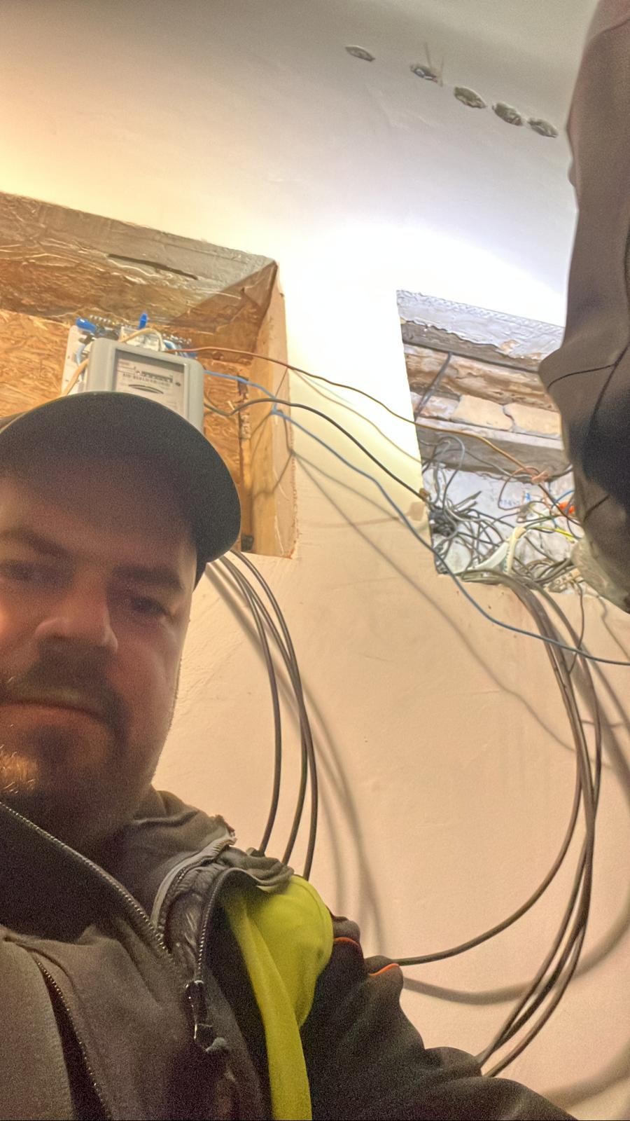Mężczyzna robiący selfie na tle rozdzielnicy elektrycznej z wystającymi kablami i fragmentem sufitu w trakcie remontu.