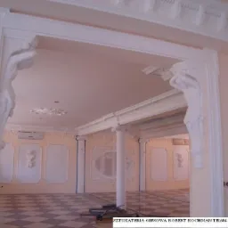 Wykończone wnętrze z dekoracyjną sztukaterią gipsową: kolumny, listwy przysufitowe i płaskorzeźby na ścianach, widok przez ozdobny portal z figurami. Podłoga w szachownicę.