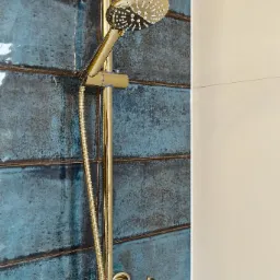 Złota bateria prysznicowa z deszczownicą na tle ściany z turkusowych płytek o nieregularnej fakturze, obok biała ściana.