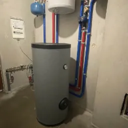 Szary bojler z podłączonymi rurami w kolorach niebieskim i czerwonym, dodatkowy biały zbiornik wyrównawczy nad nim, instalacja w surowym pomieszczeniu z betonową podłogą i ścianami.
