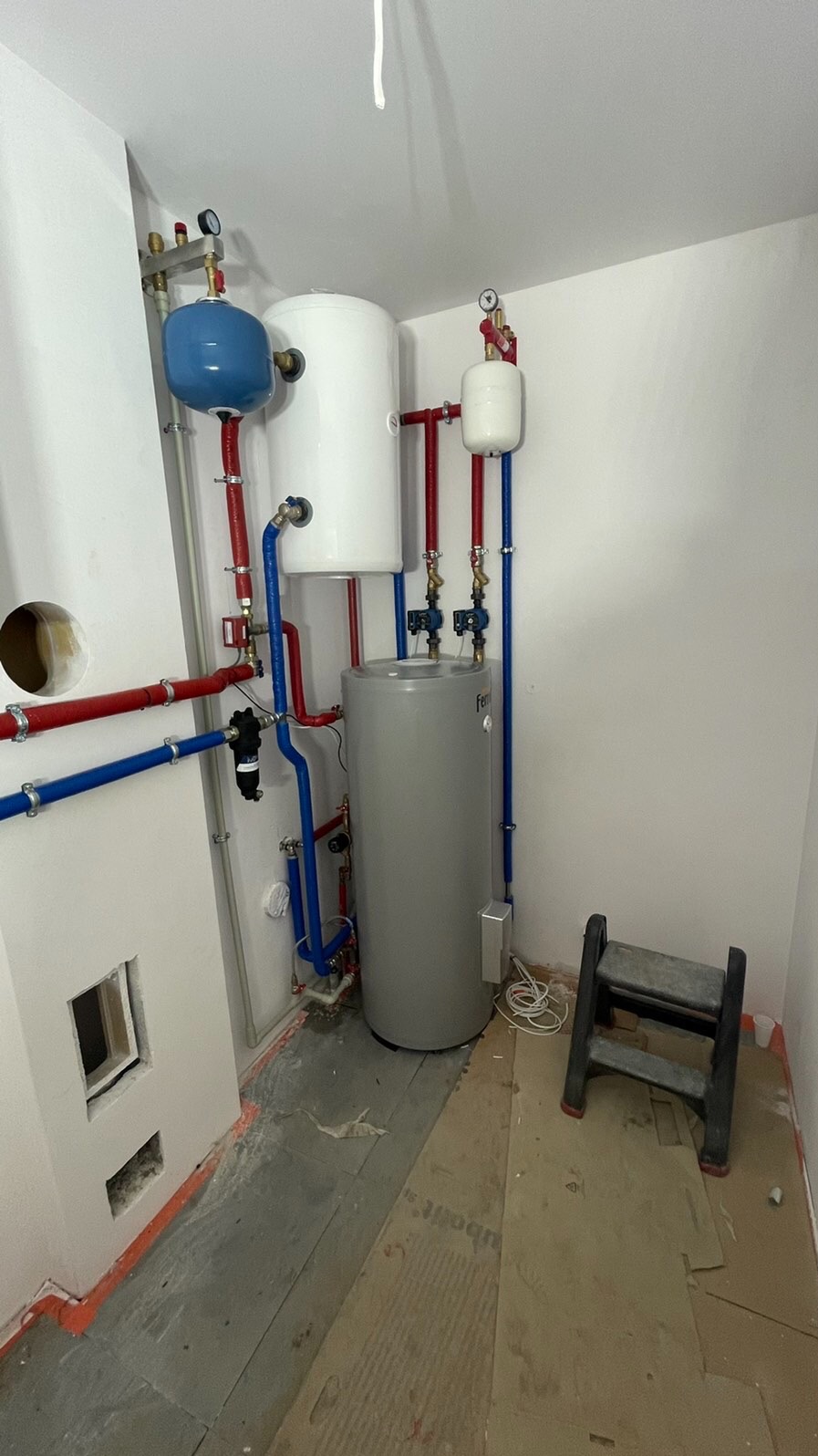 Instalacja grzewcza w kotłowni w trakcie prac wykończeniowych: szary zbiornik, biały zasobnik, niebieski zbiornik wyrównawczy, czerwone i niebieskie rury, drabinka malarska, surowa podłoga.