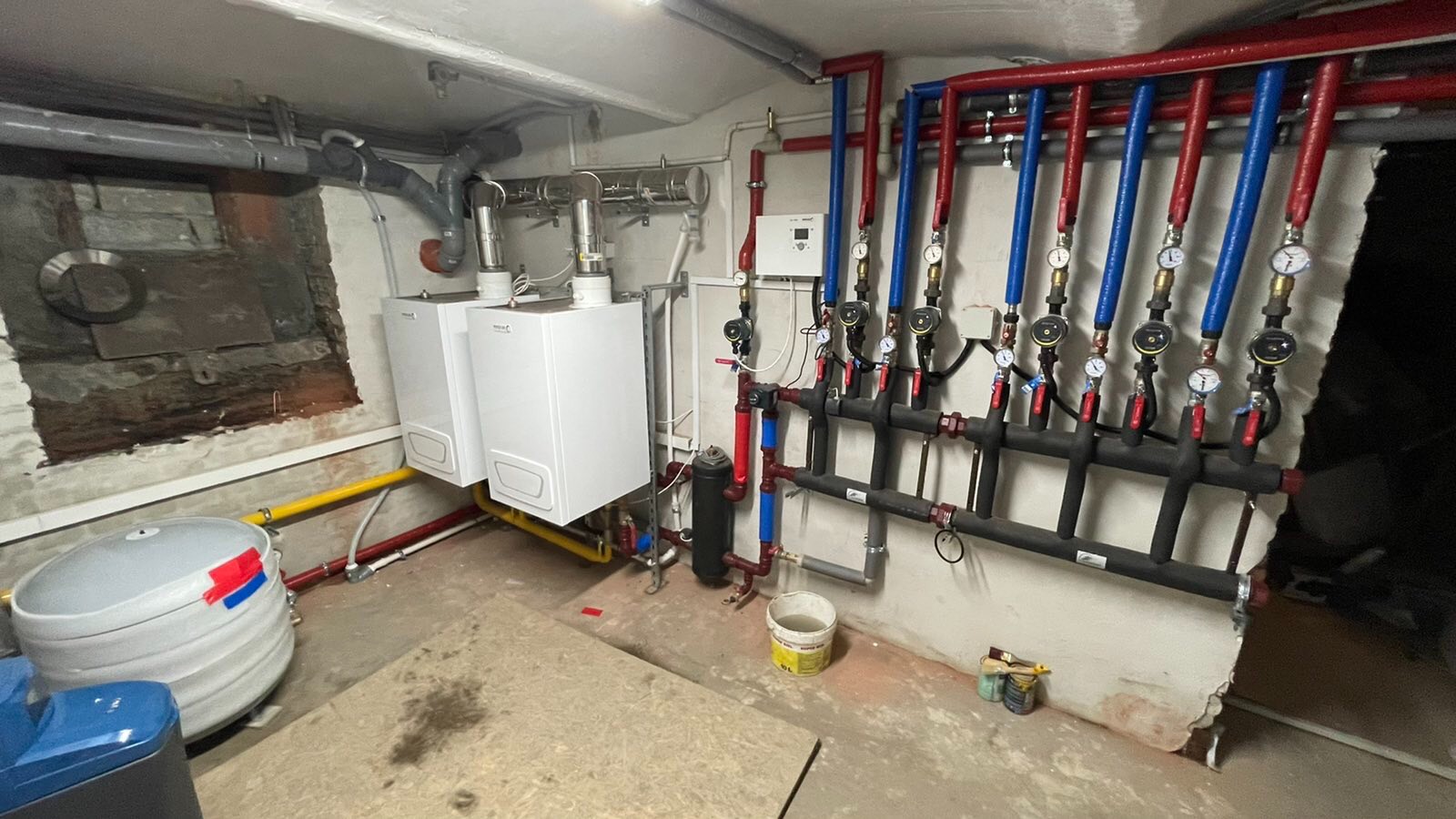 Dwa białe piece CO wiszące na ścianie w piwnicy, z widocznymi rurami w izolacji czerwonej i niebieskiej, oraz czarną instalacją rozdzielającą wodę z manometrami, na podłodze płyta OSB i wiaderko...