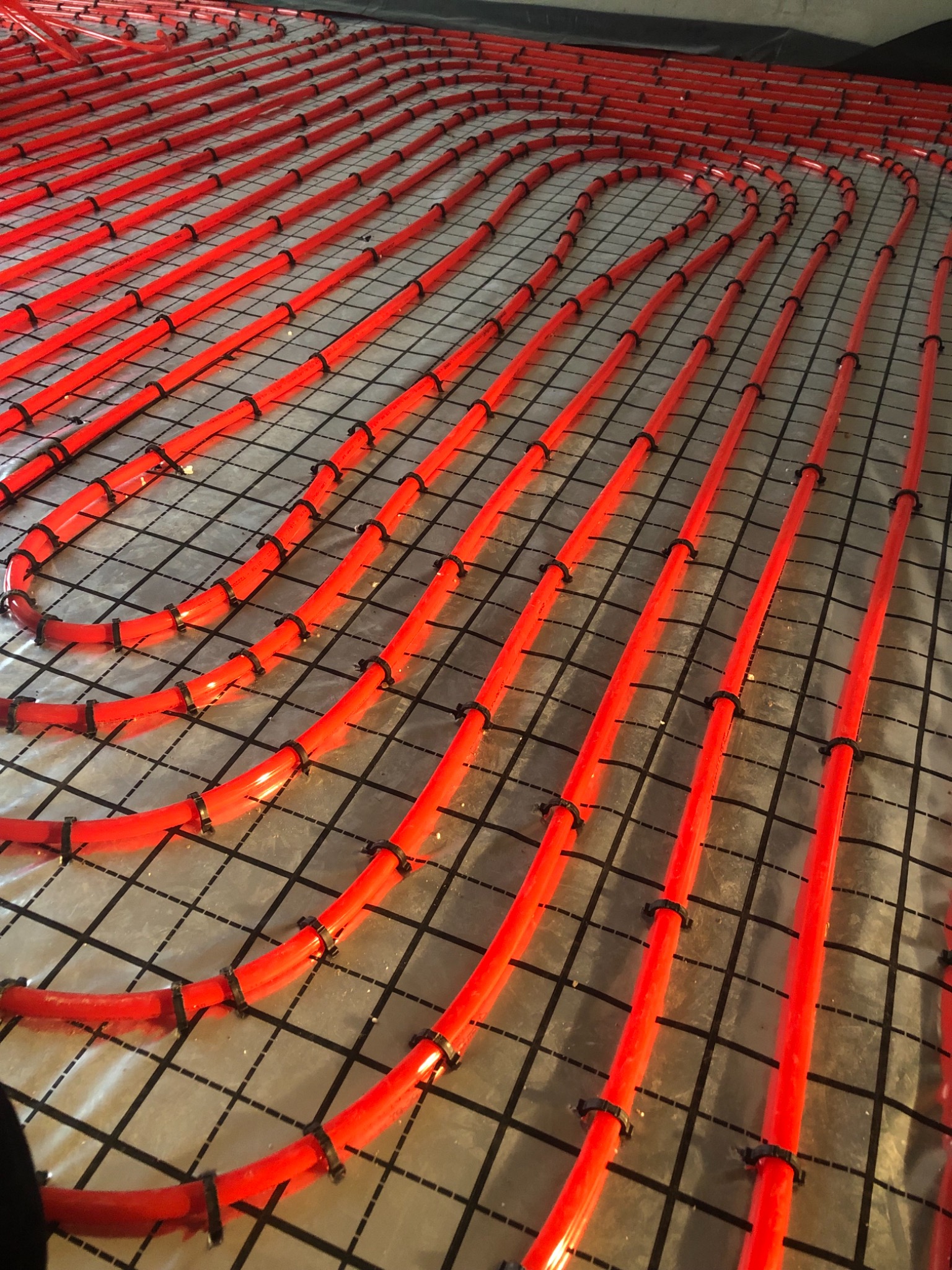 Czerwone rury ogrzewania podłogowego ułożone na siatce montażowej, widoczne czarne klipsy mocujące, przygotowanie do wylewki.
