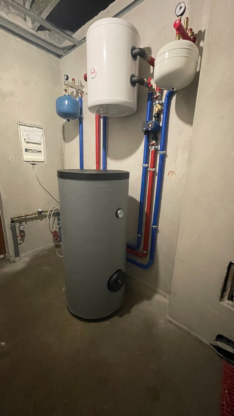 Szary bojler z podłączonymi rurami w kolorach niebieskim i czerwonym, dodatkowy biały zbiornik wyrównawczy nad nim, instalacja w surowym pomieszczeniu z betonową podłogą i ścianami.
