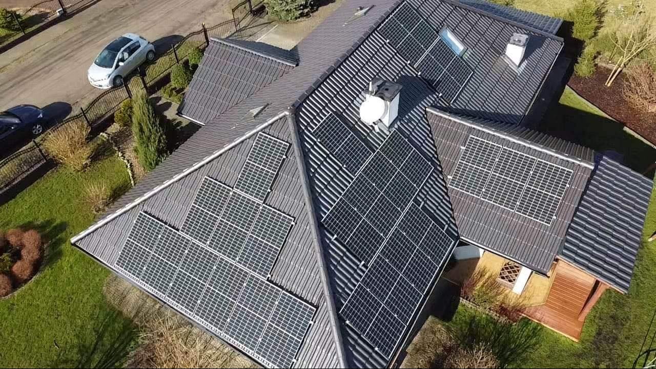 Widok z góry na dom z szarym dachem pokrytym panelami słonecznymi, z widocznymi dwoma samochodami na podjeździe i zadbanym trawnikiem.