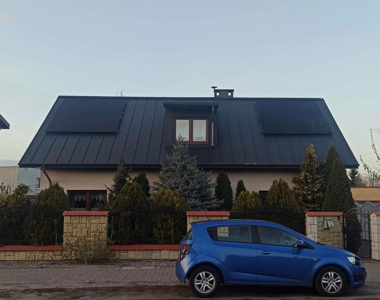 Dom jednorodzinny z ciemną blachodachówką i panelami fotowoltaicznymi na dachu, sfotografowany z poziomu ulicy, z zaparkowanym niebieskim samochodem na pierwszym planie.