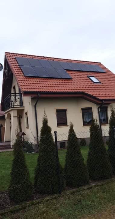 Dom jednorodzinny z czerwoną dachówką, na której zamontowane są panele słoneczne; widoczny balkon z czarną balustradą i rząd tuj wzdłuż ogrodzenia.