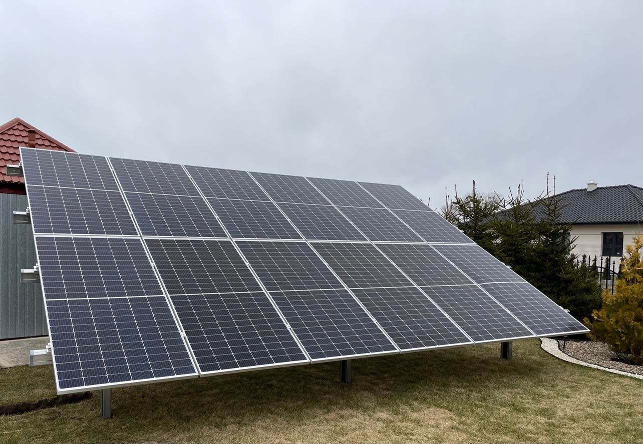 Instalacja paneli słonecznych na konstrukcji wsporczej, widok z boku na tle trawnika, domu i pochmurnego nieba.