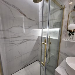 Krystian Gazowski - Nowoczesna kabina prysznicowa z przesuwnymi drzwiami, wykończona złotymi elementami, marmurowe płytki na ścianach i podłodze, deszczownica w kolorze złota, minimalistyczna umywalka.