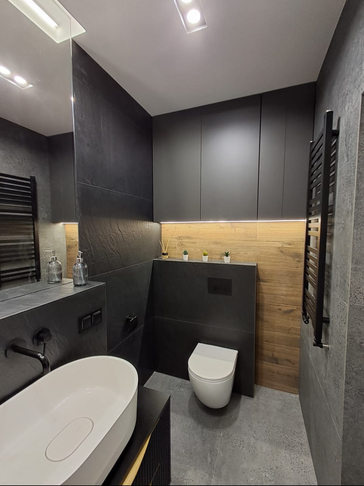 Nowoczesna łazienka z czarnymi płytkami imitującymi kamień, drewnopodobnymi panelami ściennymi, białym umywalką nablatową, wiszącą toaletą i podświetleniem LED nad szafkami.