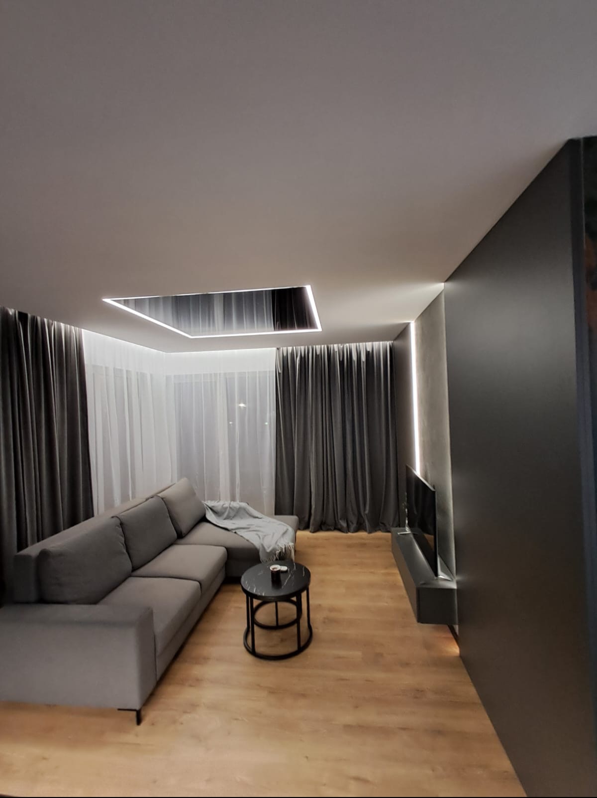 Nowoczesny salon z szarą sofą narożną, okrągłym stolikiem kawowym z czarnym blatem, drewnianą podłogą, szarymi zasłonami i podświetlanym sufitem podwieszanym.