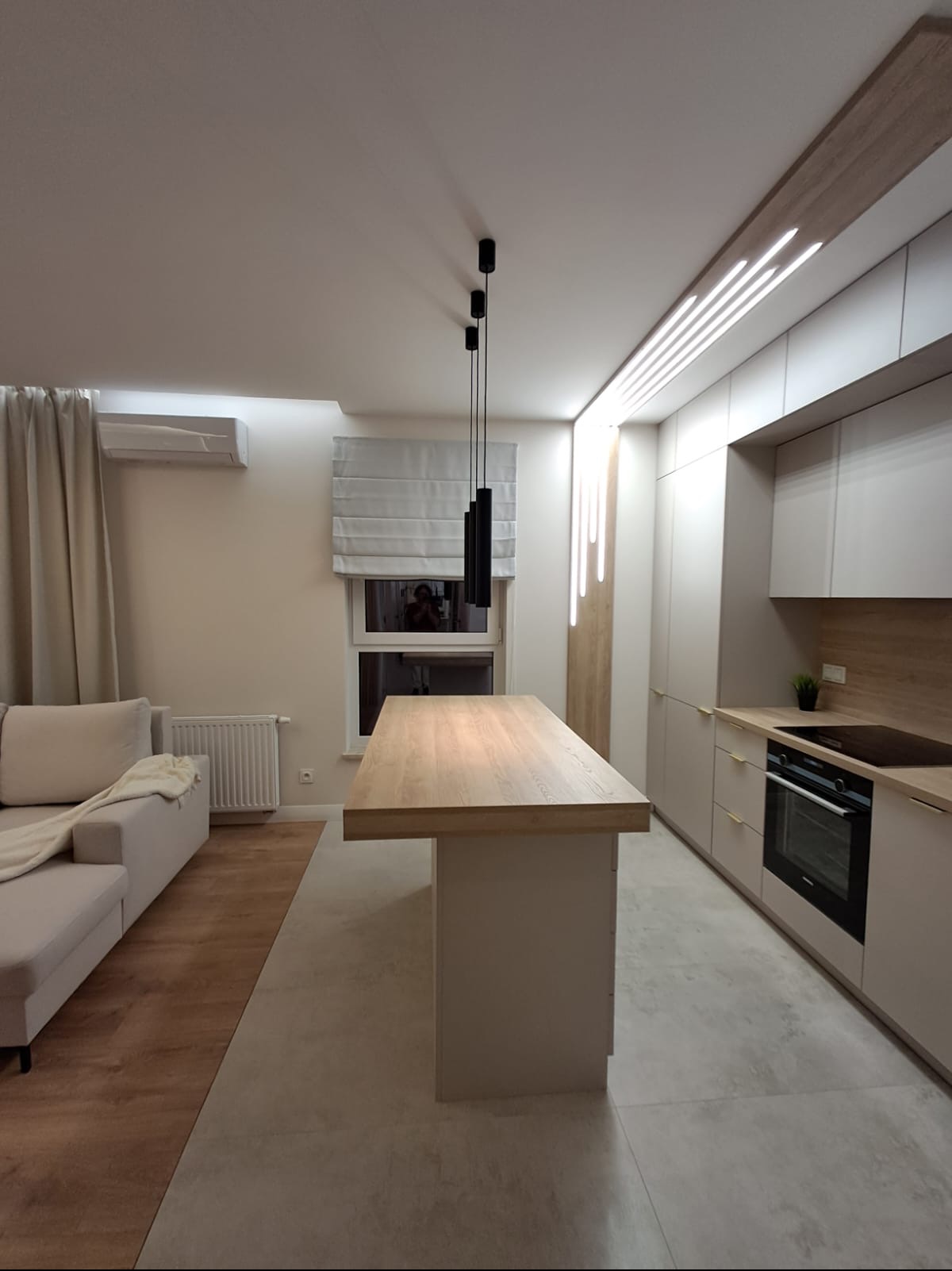 Nowoczesne wnętrze mieszkania z wyspą kuchenną o drewnianym blacie, minimalistyczne oświetlenie sufitowe i zabudowa kuchenna w jasnych odcieniach, widok na salon z sofą i zasłonami.