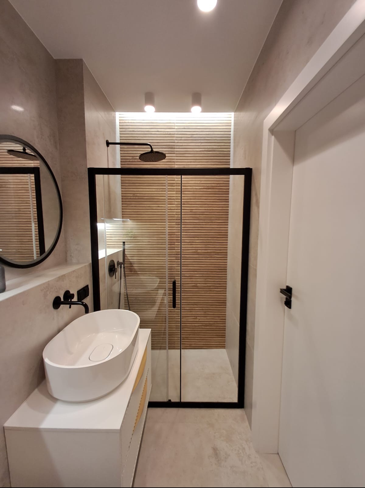 Nowoczesna łazienka z kabiną prysznicową z czarnymi profilami, drewnopodobną okładziną ścienną wewnątrz kabiny, owalną umywalką nablatową i okrągłym lustrem w czarnej ramie.
