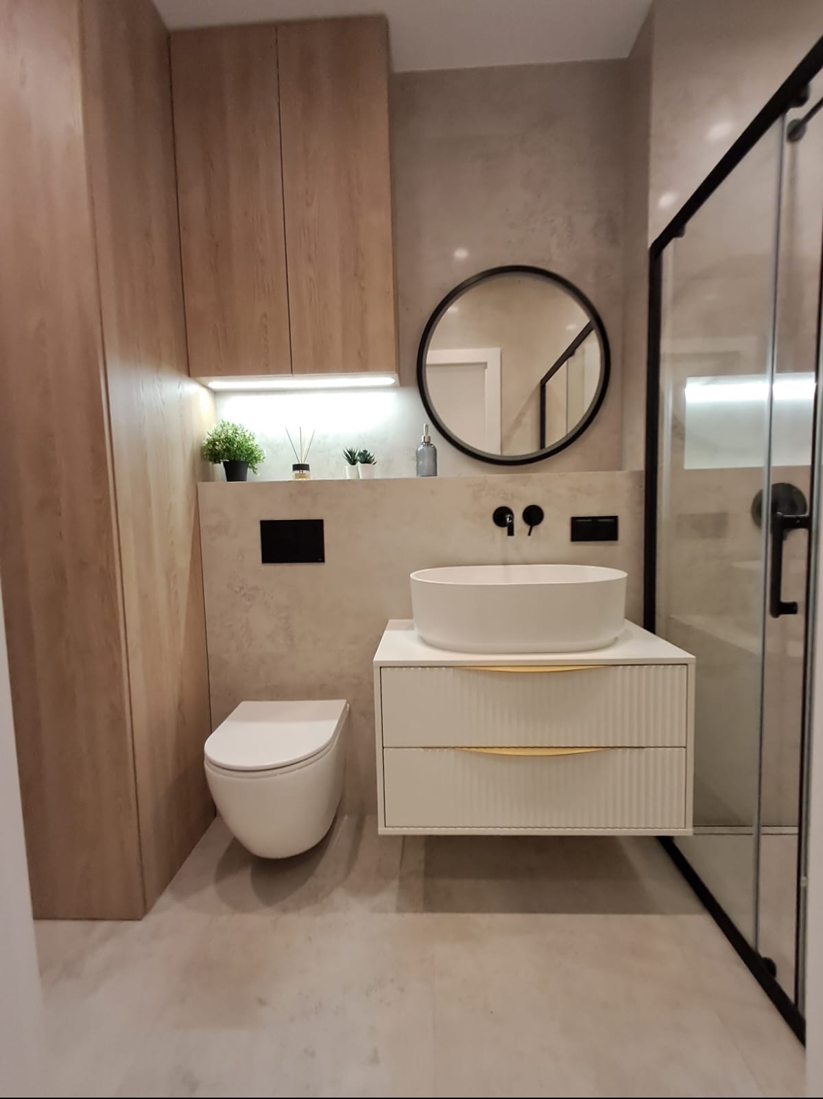 Nowoczesna łazienka z wiszącą toaletą, umywalką nablatową na szafce z frezowanymi frontami i okrągłym lustrem w czarnej ramie, oświetlona punktowo.