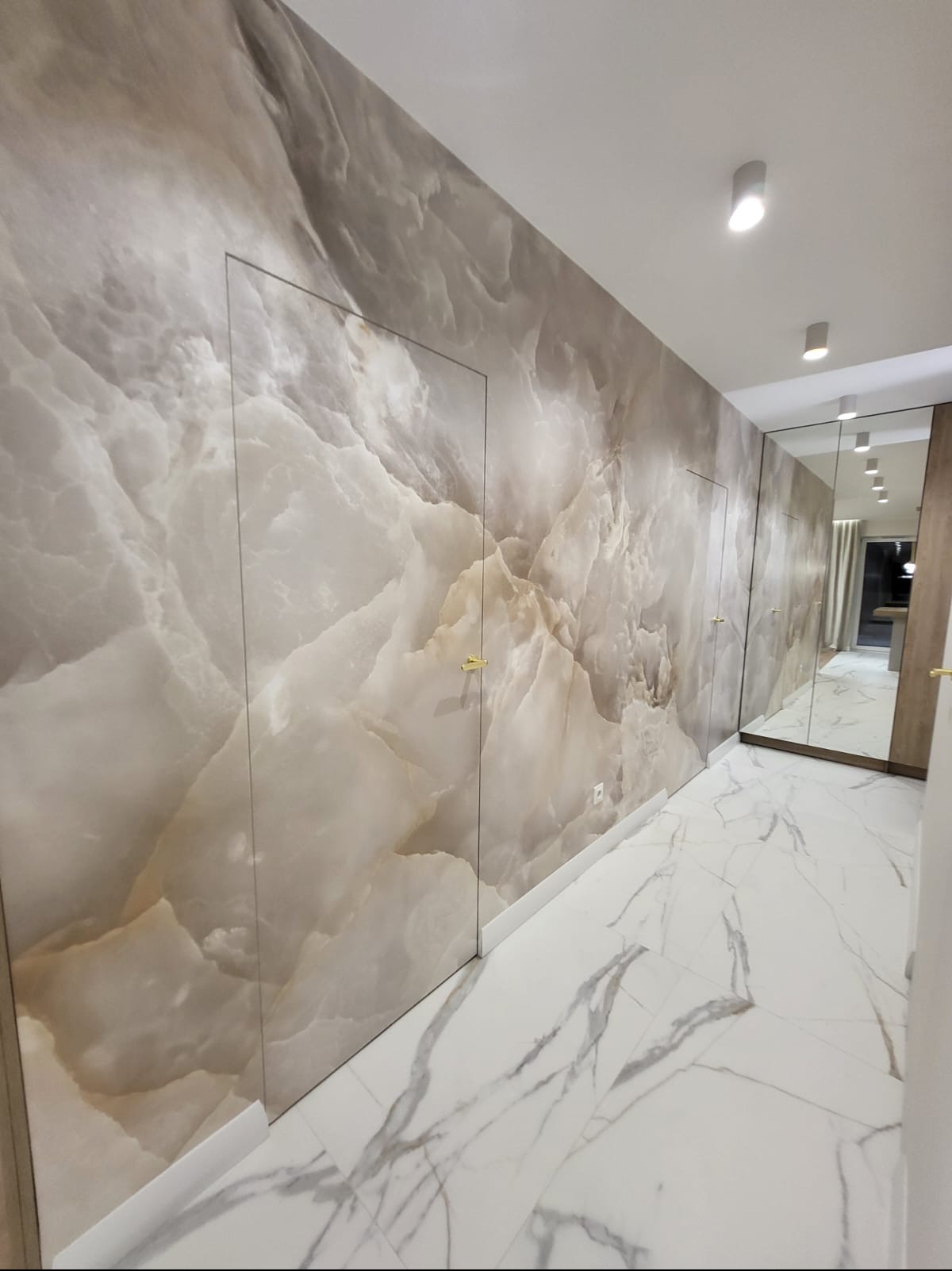 Korytarz z marmurową podłogą i ścianami imitującymi marmur z ukrytymi drzwiami zlicowanymi ze ścianą, złote klamki i lustro na końcu korytarza.