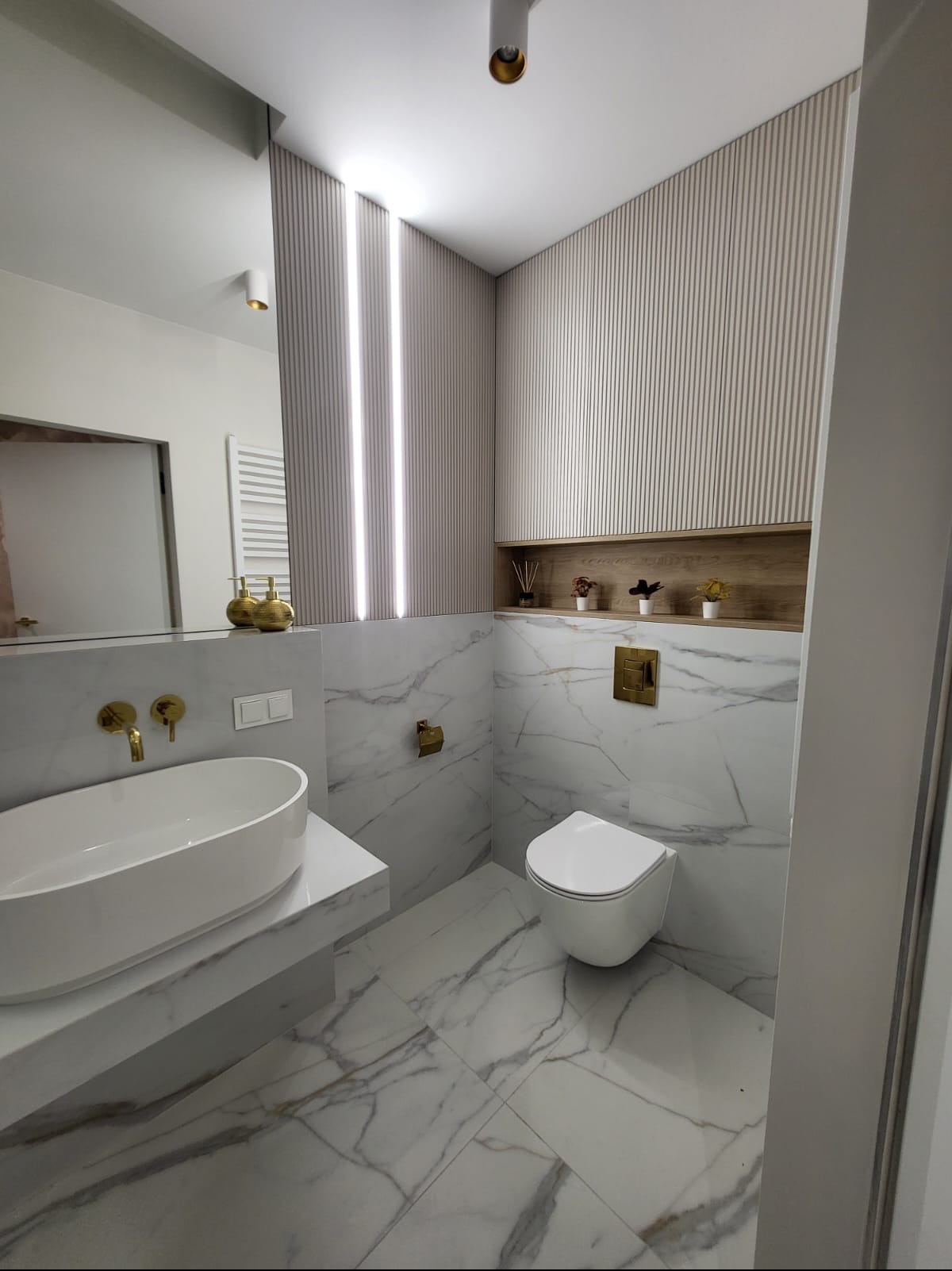 Nowoczesna łazienka z marmurowymi płytkami, owalną umywalką nablatową, złotą armaturą i podświetlanymi panelami ściennymi.