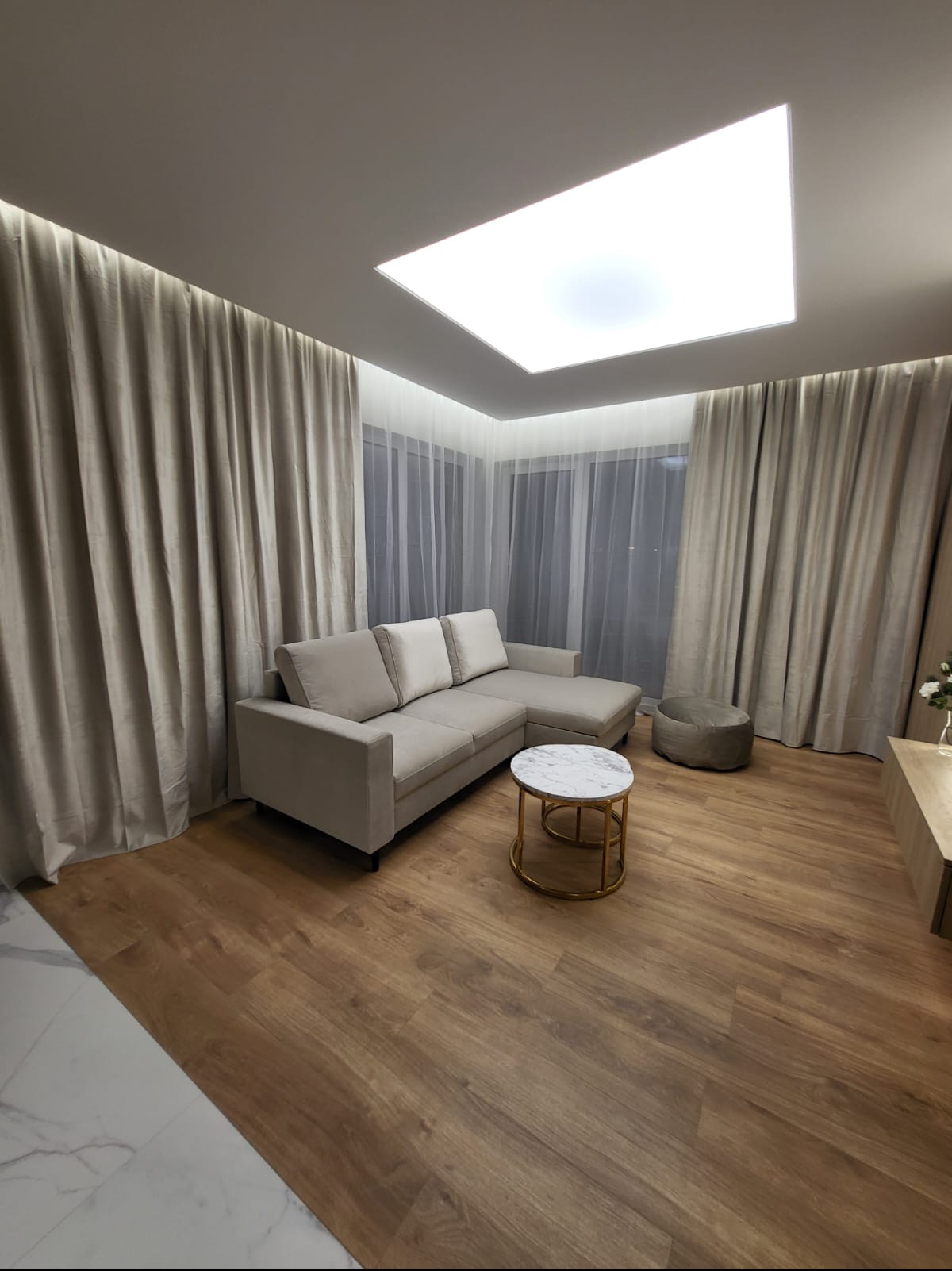 Salon z drewnopodobną podłogą, beżową sofą narożną, okrągłym stolikiem z marmurowym blatem i jasnymi zasłonami, oświetlony kwadratowym panelem LED na suficie.