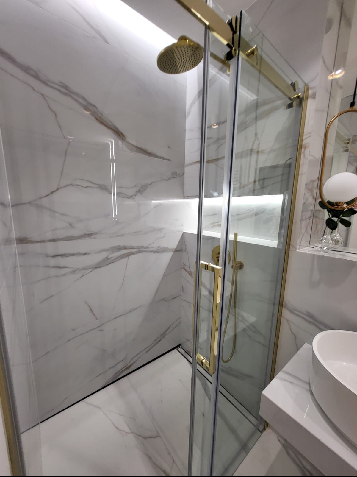 Nowoczesna kabina prysznicowa z przesuwnymi drzwiami, wykończona złotymi elementami, marmurowe płytki na ścianach i podłodze, deszczownica w kolorze złota, minimalistyczna umywalka.