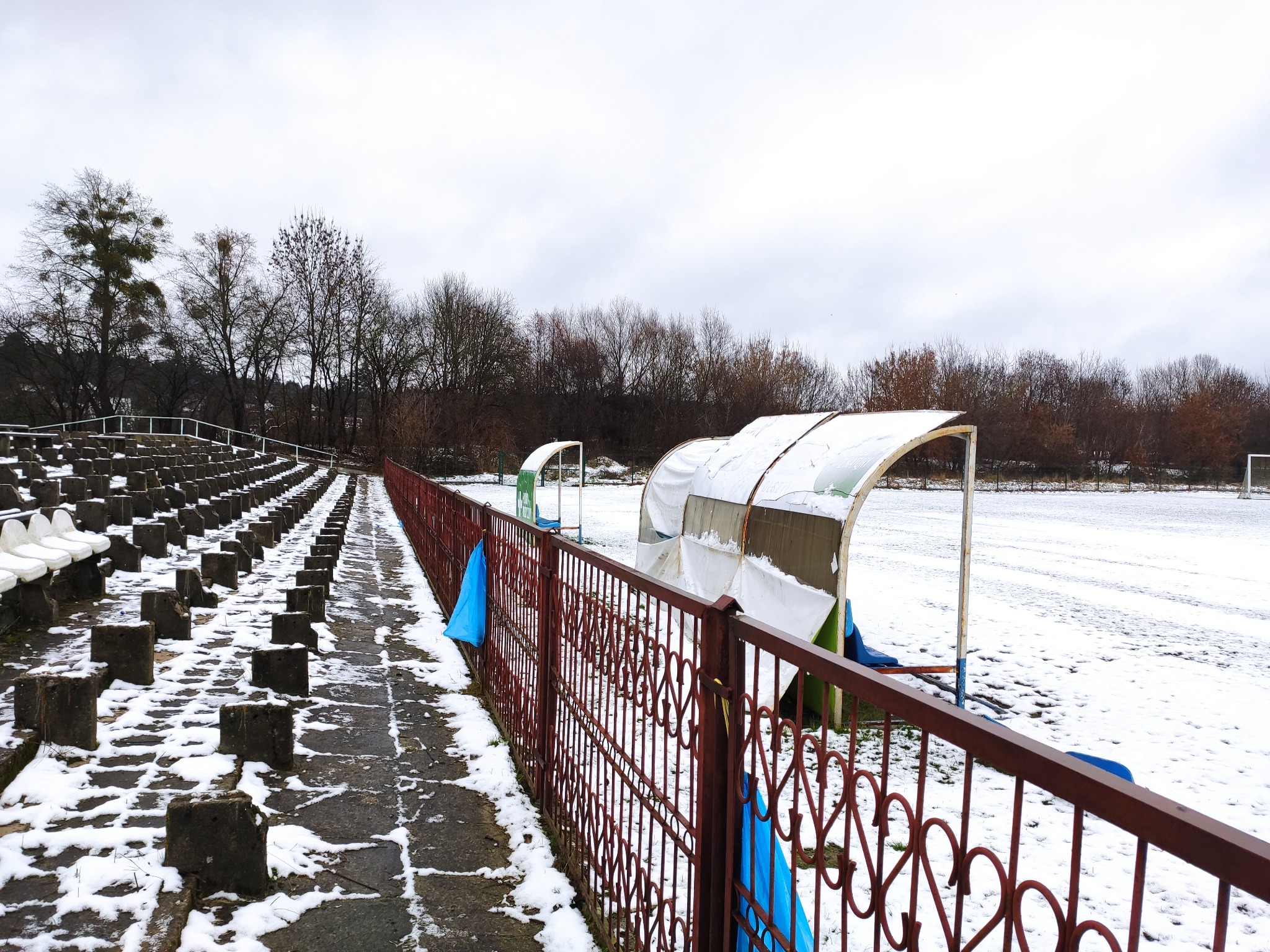 Zimowy krajobraz stadionu sportowego z trybunami, zadaszonymi ławkami rezerwowych i boiskiem pokrytym śniegiem, widzianymi przez ozdobne, metalowe ogrodzenie.
