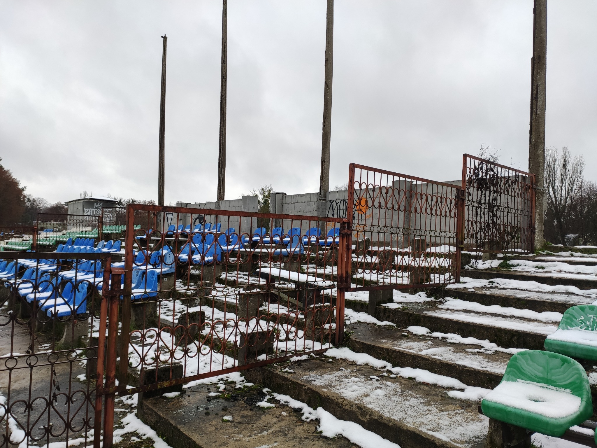 Zewnętrzne trybuny stadionu sportowego, z niebieskimi i zielonymi siedzeniami pokrytymi śniegiem, ogrodzone rdzewiejącą metalową barierką z motywem serca, w tle widoczne maszty oświetleniowe...