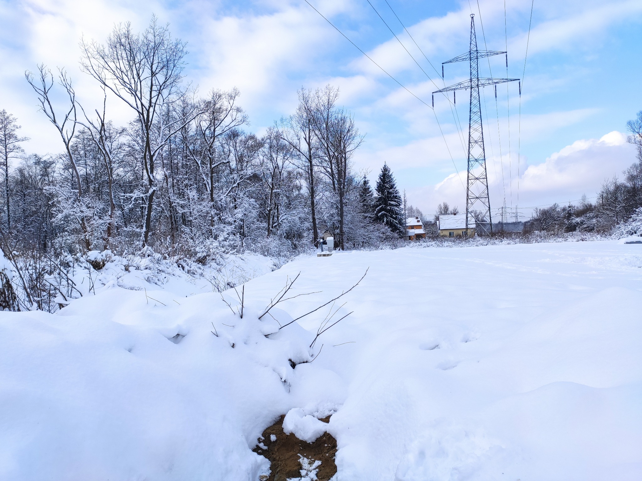 Zimowy krajobraz z linią wysokiego napięcia i zasypanym śniegiem terenem, w tle budynek mieszkalny.