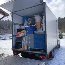 Otwarty tył białego furgonetki załadowanego różnymi przedmiotami, w tym kołem samochodowym, skrzyniami, meblami i sprzętem AGD, stojącego na śniegu obok torów kolejowych.