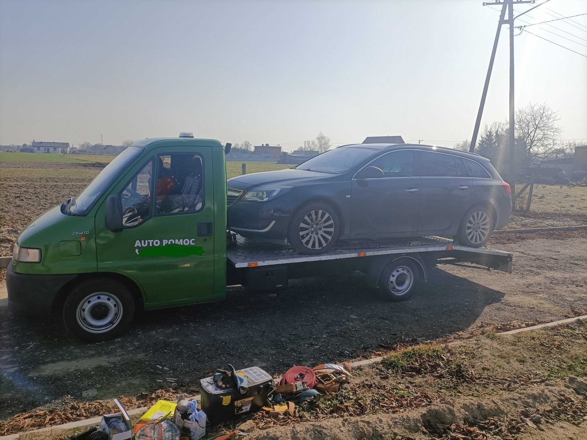 Zielona laweta z napisem 'Auto Pomoc' przewozi ciemny samochód osobowy na tle pola i słupów energetycznych.