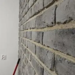Ściana wykończona szarą cegłą klinkierową z jasną fugą, widoczny fragment białej ściany z gniazdkiem elektrycznym i czerwony uchwyt narzędzia w dolnym rogu.